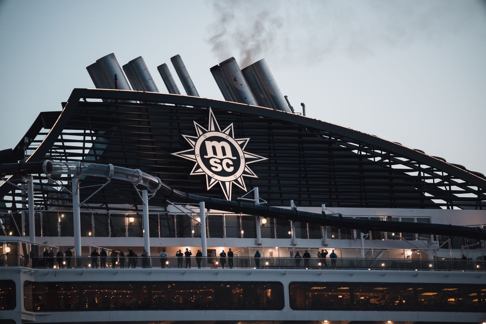 Guests on the upper decks of an MSC Cruises ship at dusk beneath its funnels