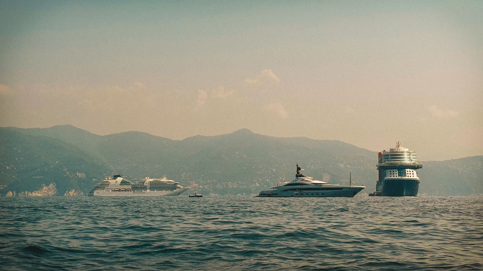 Ships and yachts at anchor on a hazy sea beneath a mountain coast